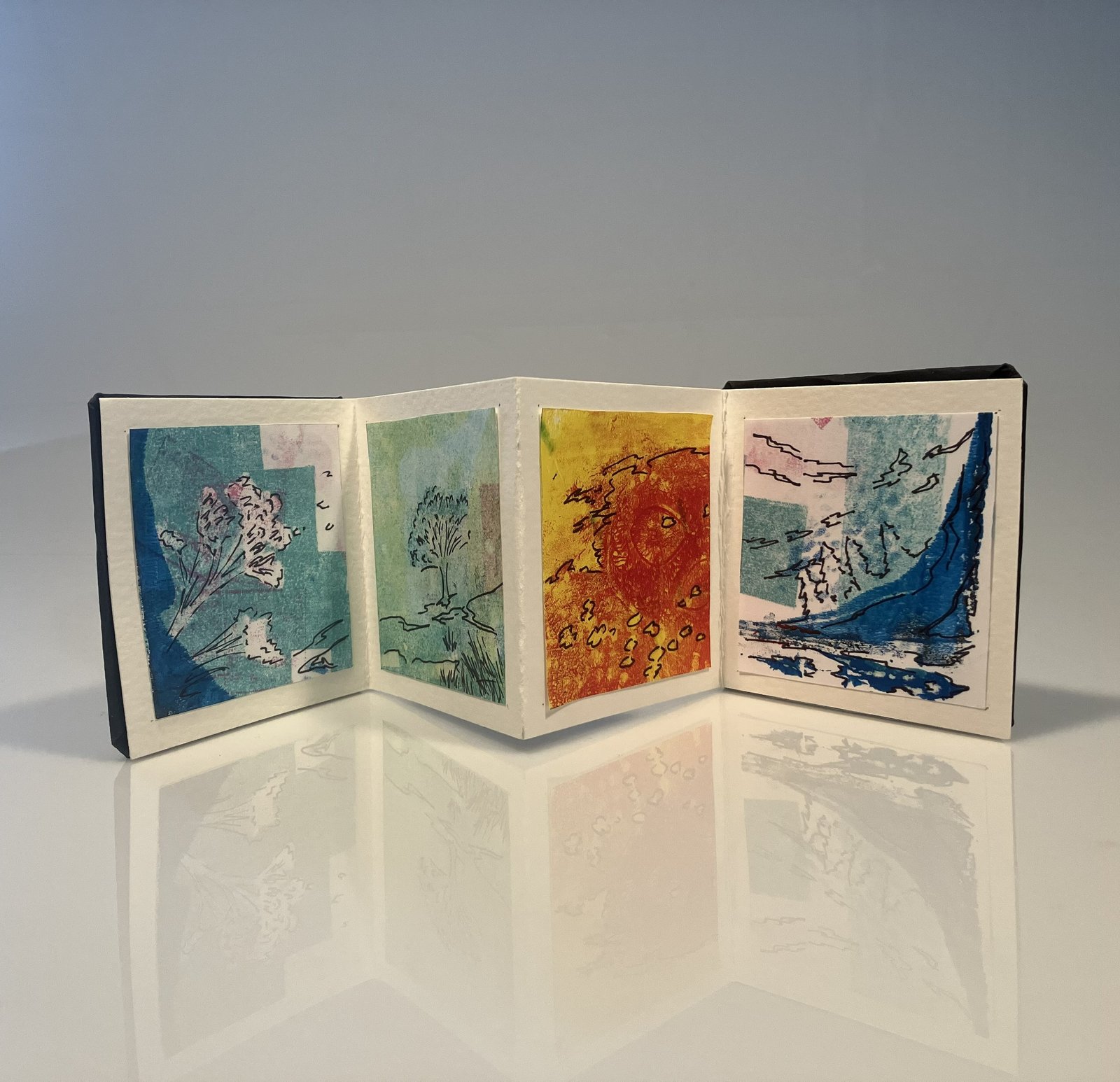 An accordion book with depictions of the four seasons drawn onto each of the main panels. The book is set against a reflective white background with overhead lighting.
